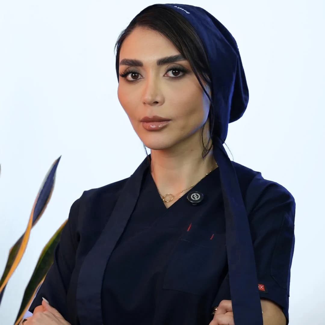 د. سارا أرجانج بزي الجراحة الكحلي وقبعة الجراحة، وذراعاها متشابكتان، مصوّرة أمام خلفية فاتحة سادة.