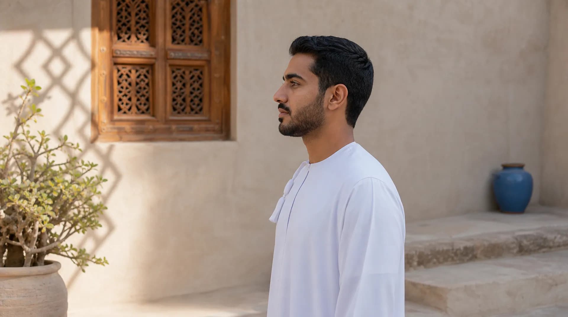 رجل عُماني بوضع جانبي طبيعي أمام جدار جصّي فاتح في ضوء جانبي ناعم.
